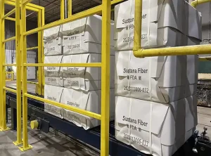 Große Produktwürfel, in weißes Papier eingewickelt, auf einem Förderband. Die Würfel sind mit schwarzer Tinte mit QR-Codes und alphanumerischen Zeichen beschriftet.