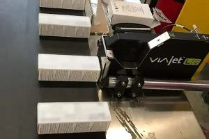 Matthews Marking Systems VIAjet L12-Thermo Tinte -Drucker, der weiße Produktkartons während ihres Transports auf einem Produktionsförderband bedruckt.