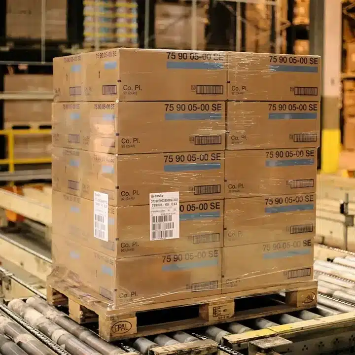 Auf einer Holzpalette gestapelte und mit Barcode-Etiketten versehene Kartons, die in einem Lager auf einem Fördersystem transportiert werden.