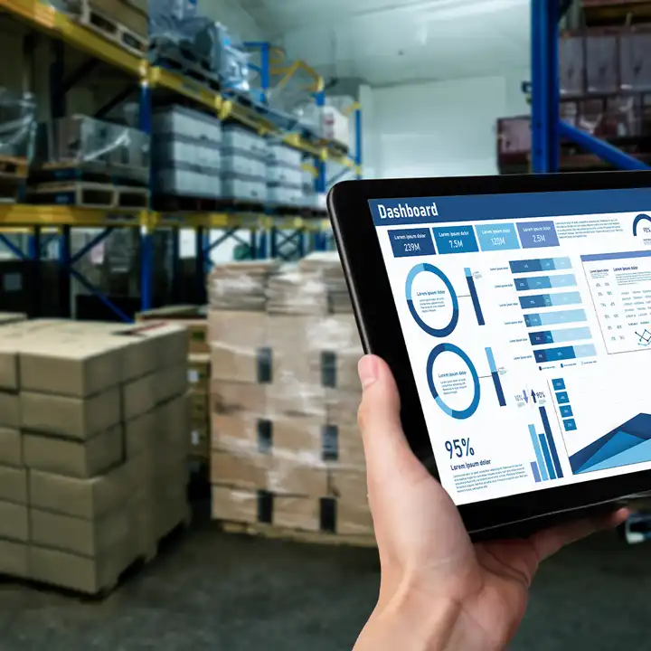 Eine Person, die ein Tablet in der Hand hält, auf dem Produktionsanalysen und Leistungsdiagramme angezeigt werden, in einem Lager mit palettiertem Lagerbestand und Lagerregalen.