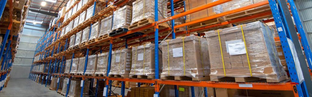 Reihen von in Schrumpffolie verpackten Paletten mit Kartons, die in industriellen Lagerregalen für den Versand und Vertrieb gelagert werden.